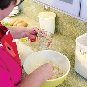 A woman mixing ingredients in a yellow bowl on a kitchen counter with a measuring cup, flour container, and potatoes, in the background. Photo: Nate Yonamine